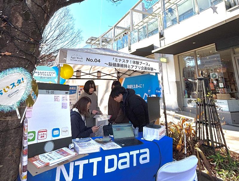写真：NTT DATAの屋外ブースでスタッフが資料を並べ、体験イベントを案内している